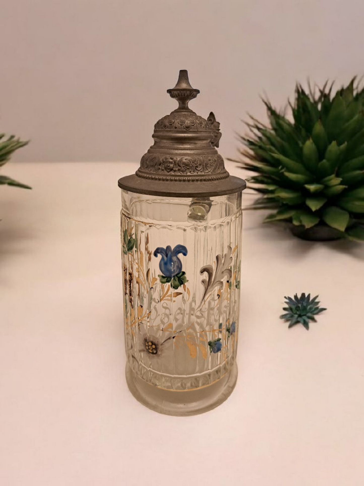 Această halbă de bere vintage, fabricată în Germania de Vest între anii 1950-1970, este o piesă de colecție remarcabilă. Realizată din sticlă pictată manual și dotată cu un capac din cositor, halba impresionează prin detaliile sale complexe și autenticitatea designului.